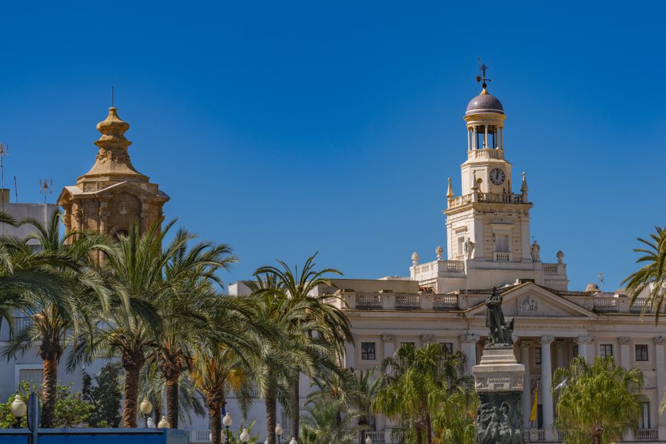 Ayuntamiento de Cádiz con arquitectura icónica y estatua, enmarcado por palmeras bajo un cielo azul claro.