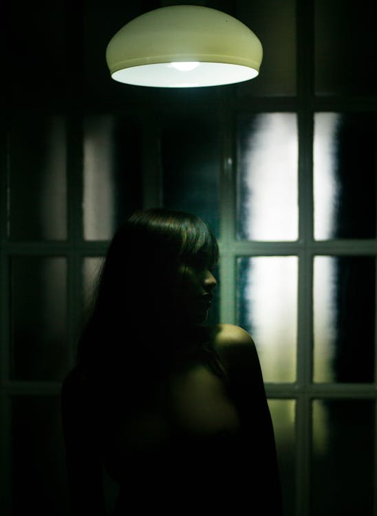 Free Emotionless brunette standing in shadows under dim light lamp in dark room against glass wall with glowing lights reflections and turning head to left Stock Photo