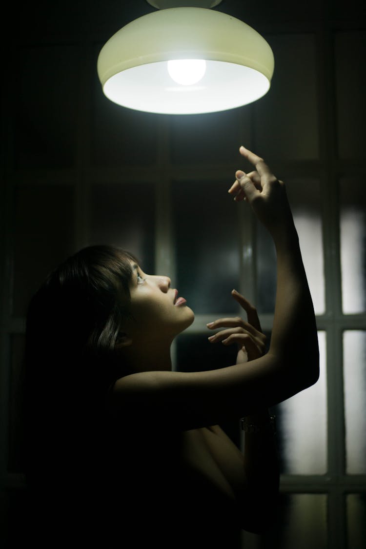 Gorgeous Woman Raising Arms To Electric Lamp In Dark Room