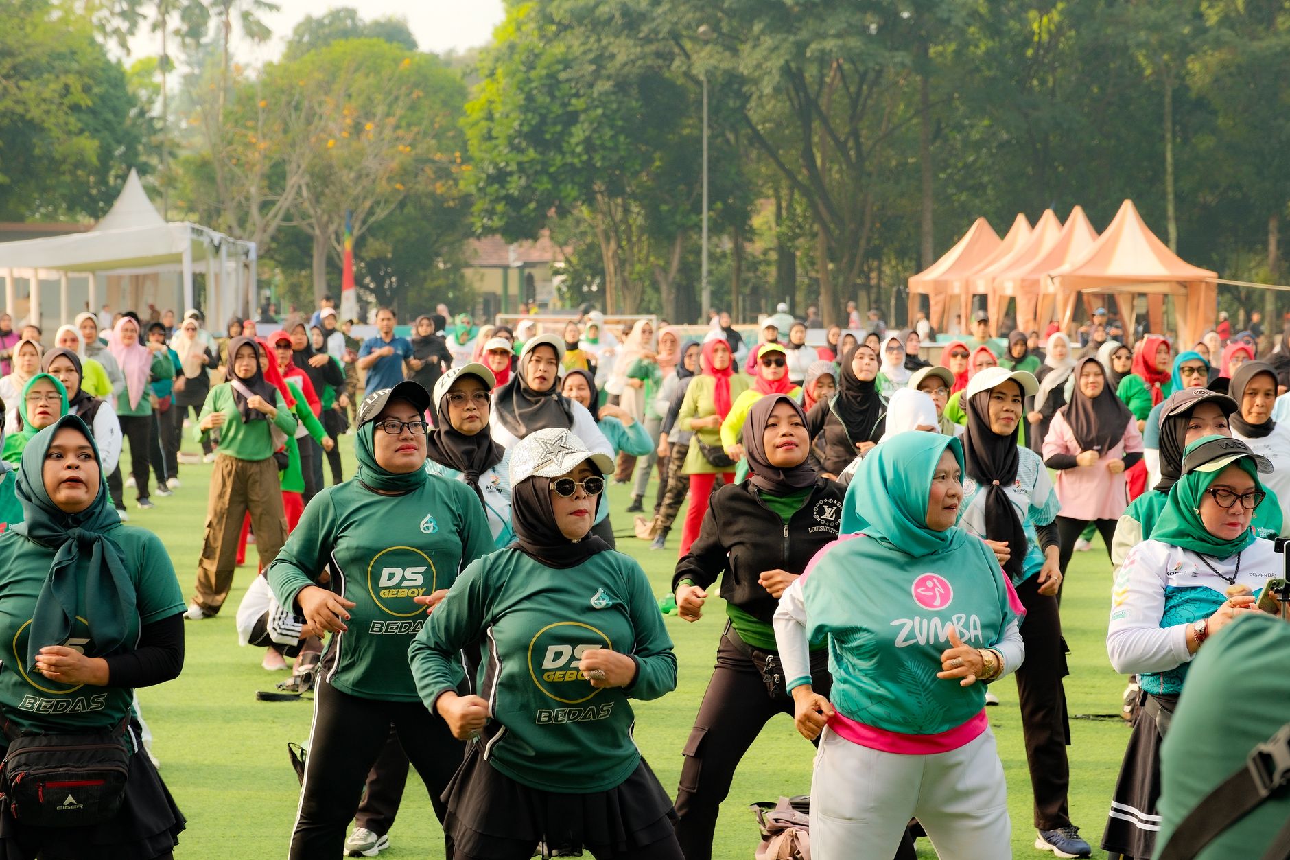 A large outdoor gathering of women participating in a Zumba class with trees and tents in the background.