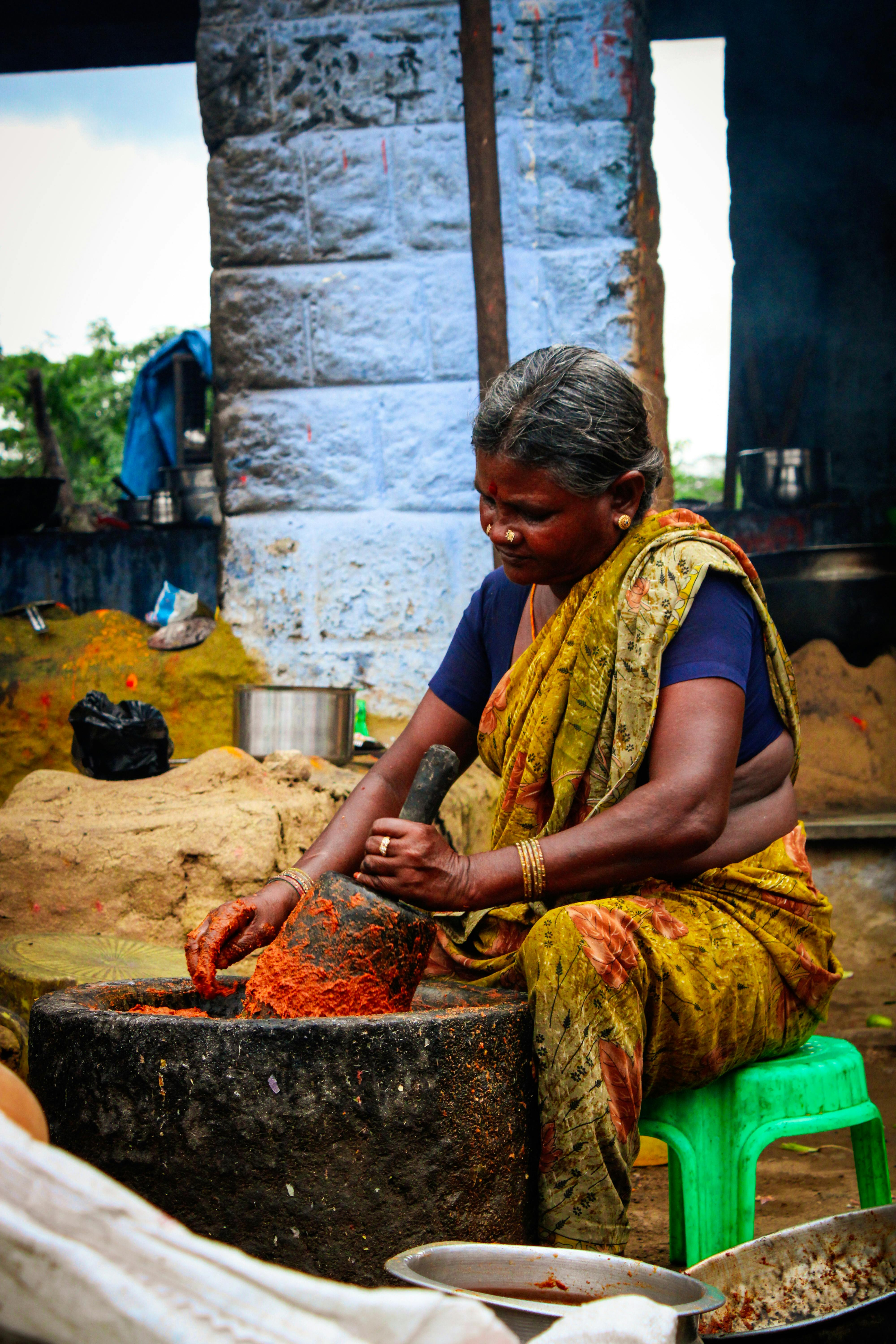 Full body senior ethnic female in colorful traditional clothes sitting on plastic stool in messy courtyard and powdering spices in big stone bowl