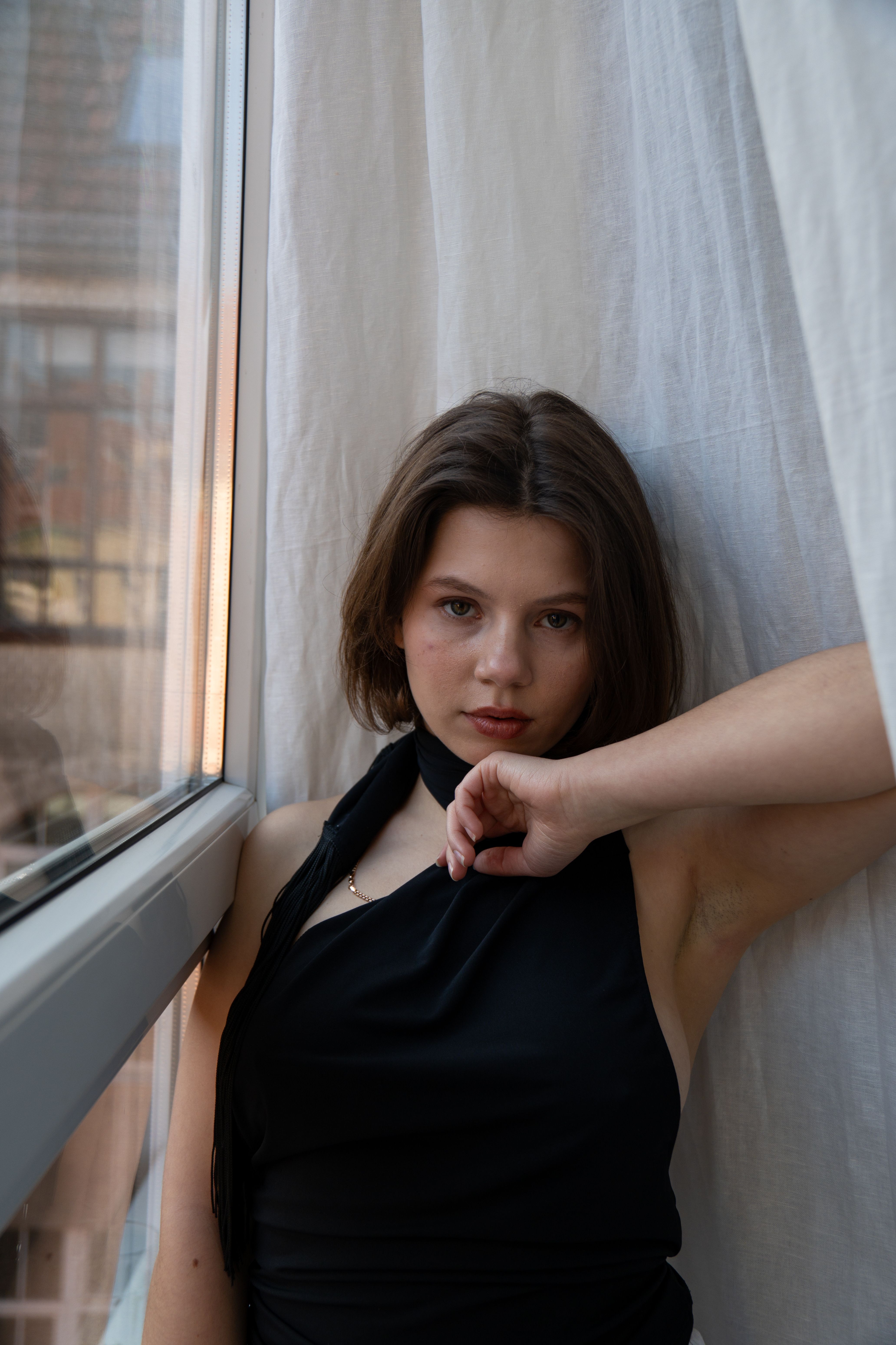 A young woman in a black dress poses by a window, exuding elegance and confidence.