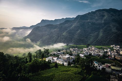 Free A serene village nestled in lush green mountains with morning mist drifting over the landscape. Stock Photo