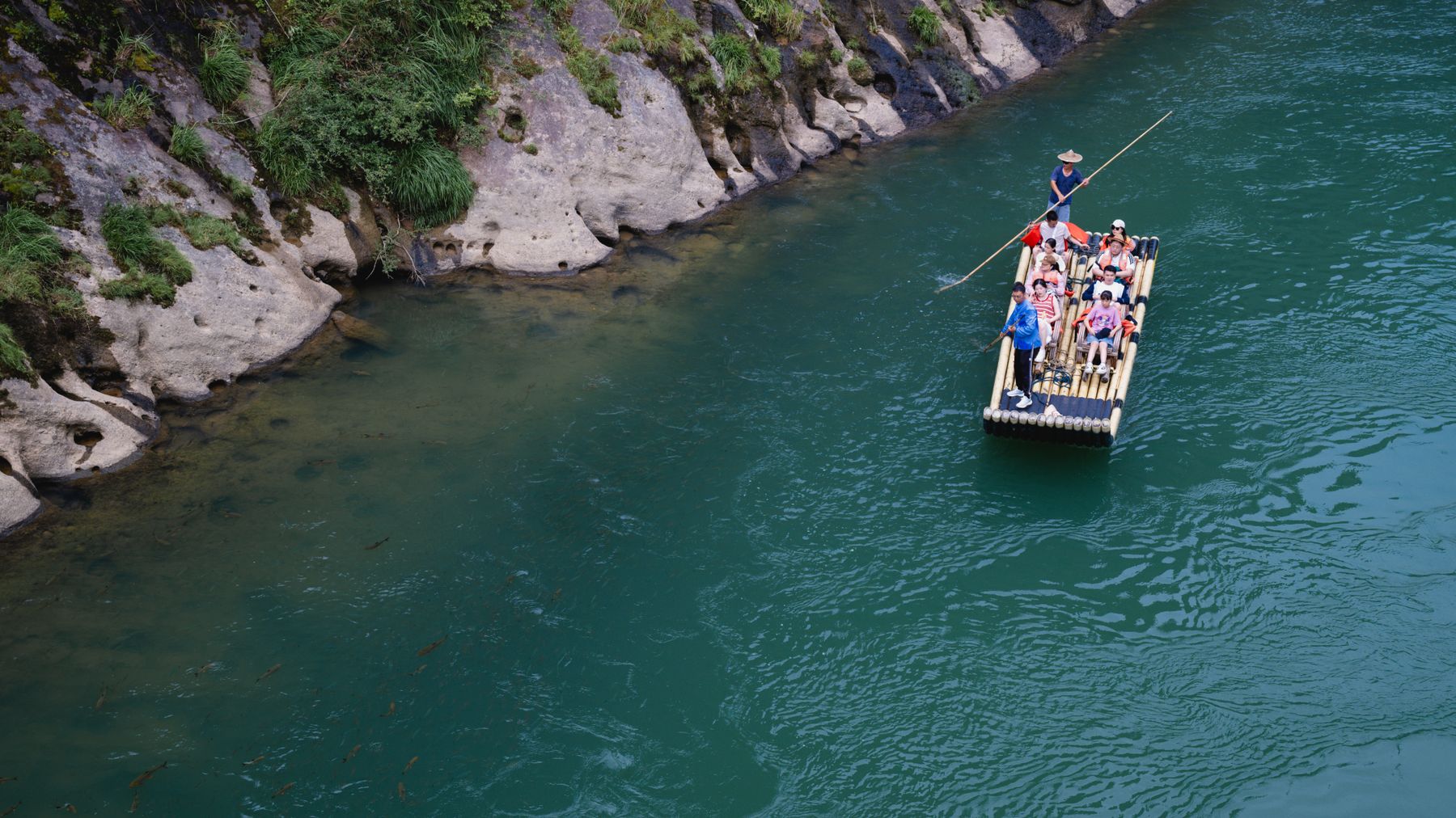 A group enjoying a serene bamboo raft ride on a turquoise river beside lush green cliffs.