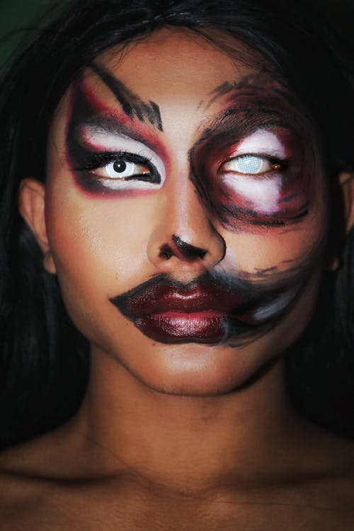 Portrait of unemotional young ethnic female with dramatic painting on face and white lens in left eye looking at camera