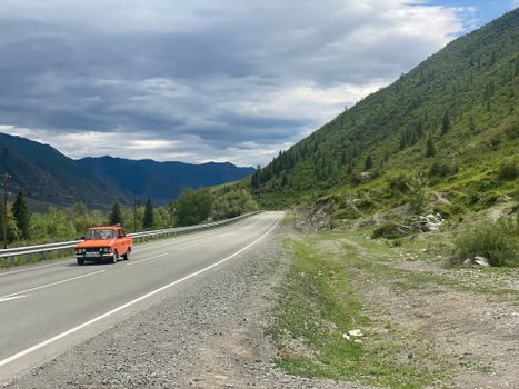 A red car travels along a scenic mountain road in the Altai Republic, Russia under a cloudy sky.