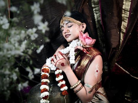 Serene Indian woman in traditional attire holding lotus, adorned with jewelry and garlands.