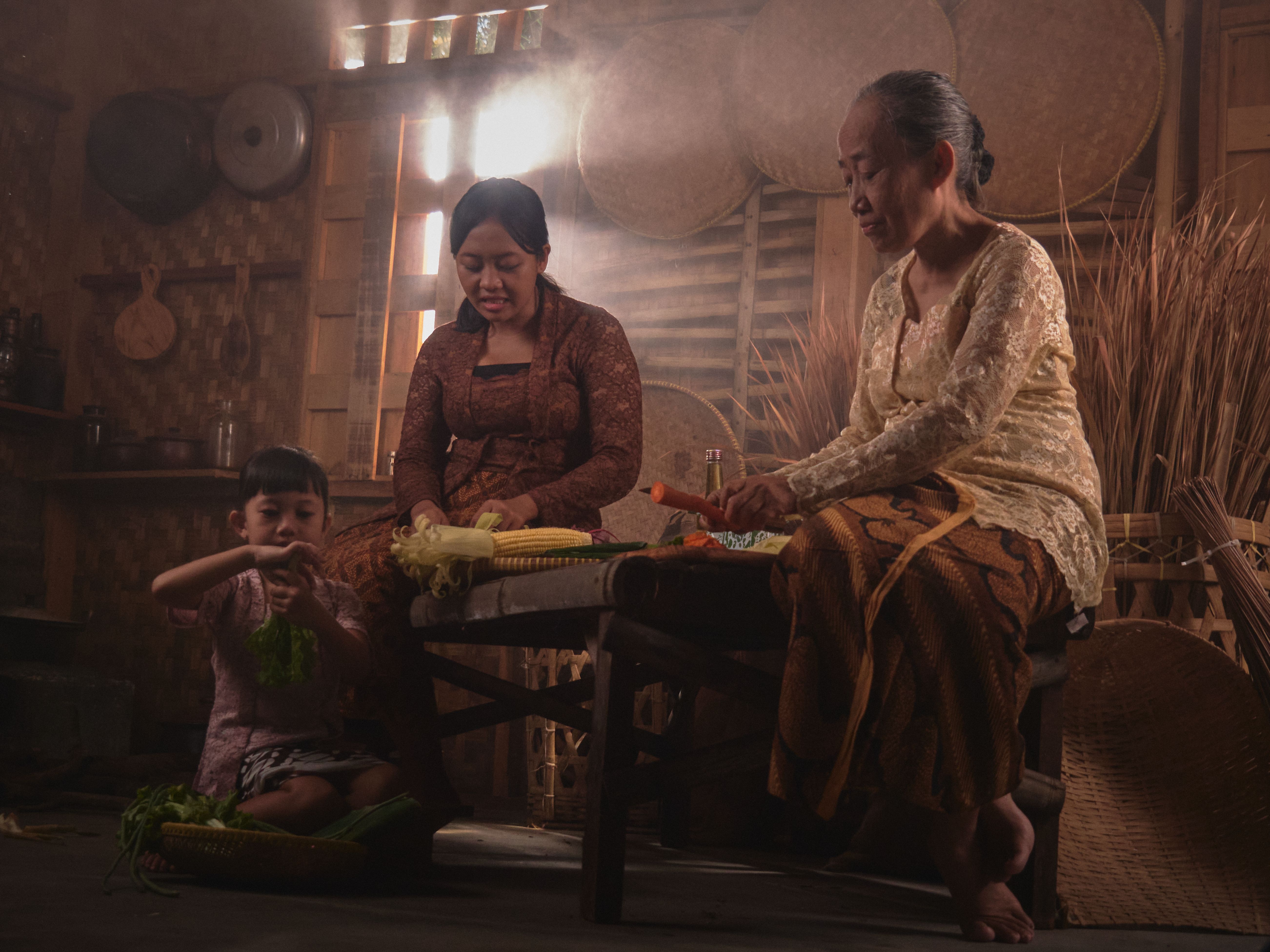 A family engages in traditional cooking in a warm, rustic Indonesian kitchen setting.
