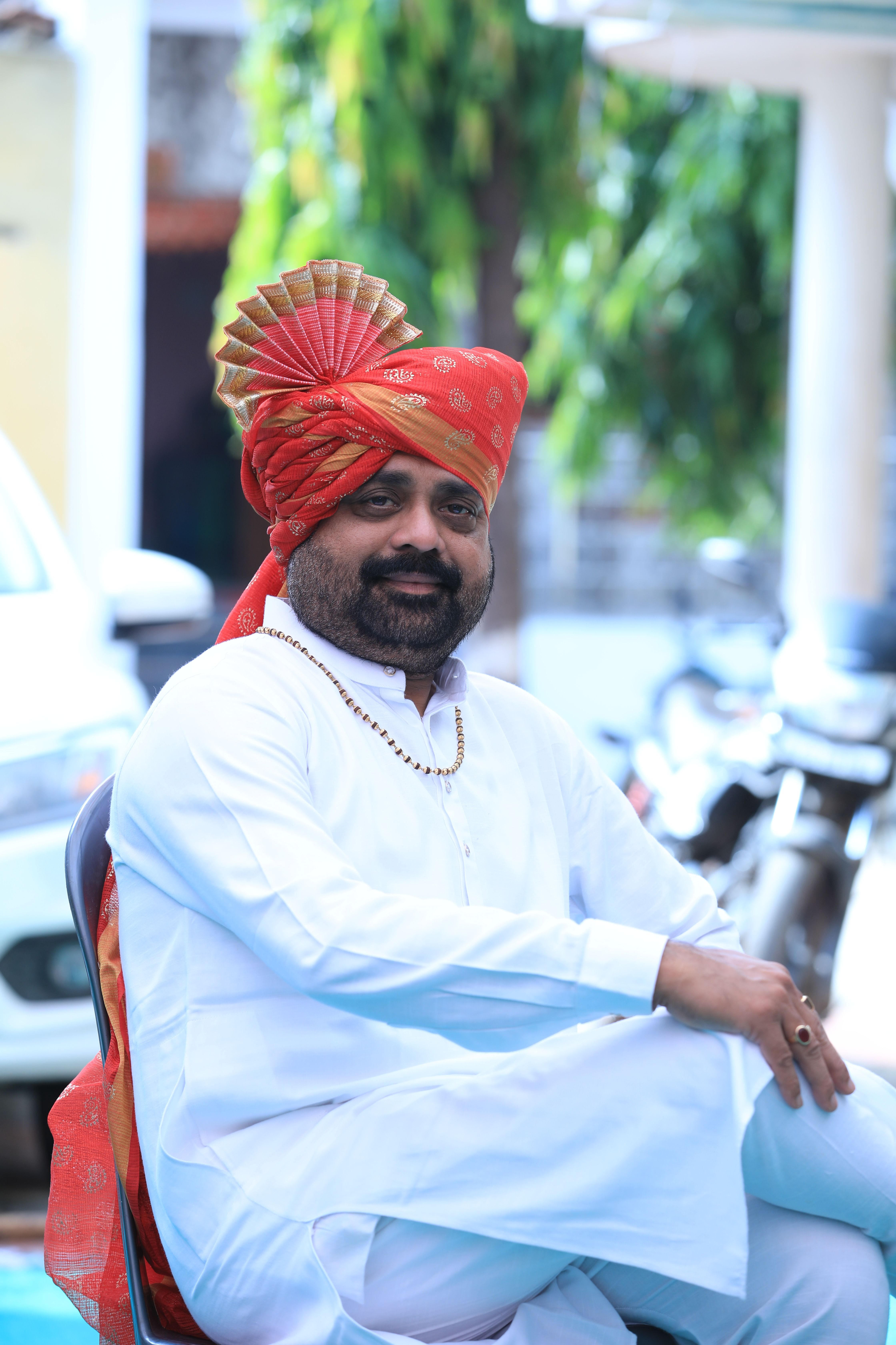 A man wearing traditional Indian attire with a vibrant turban sits outdoors during the day.