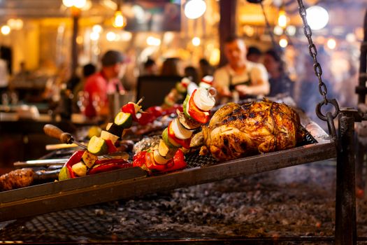 Grilled chicken and vegetable skewers over an open flame in a lively market setting.