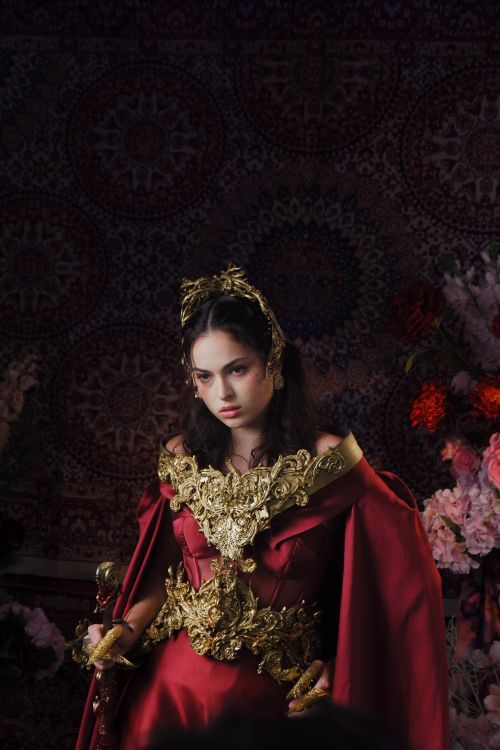 Free A young woman in a red and gold ornate dress with floral background, exuding a regal presence. Stock Photo