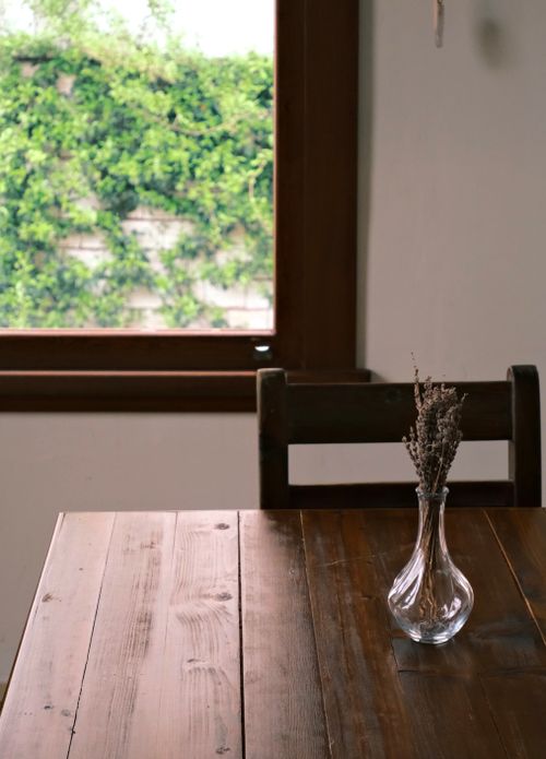 Free A rustic wooden table with a glass vase and dried flowers by a window in a cozy room. Stock Photo