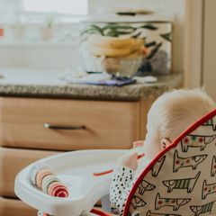 Fair-haired baby seen from behind in a white folding high chair with a red-trimmed animal-print seat pad, in a kitchen