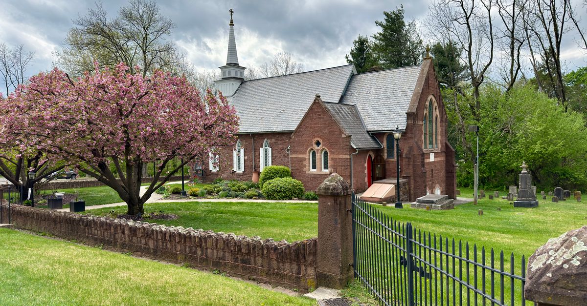 A picturesque red brick church surrounded by lush greenery and a blossoming cherry tree under a cloudy sky.