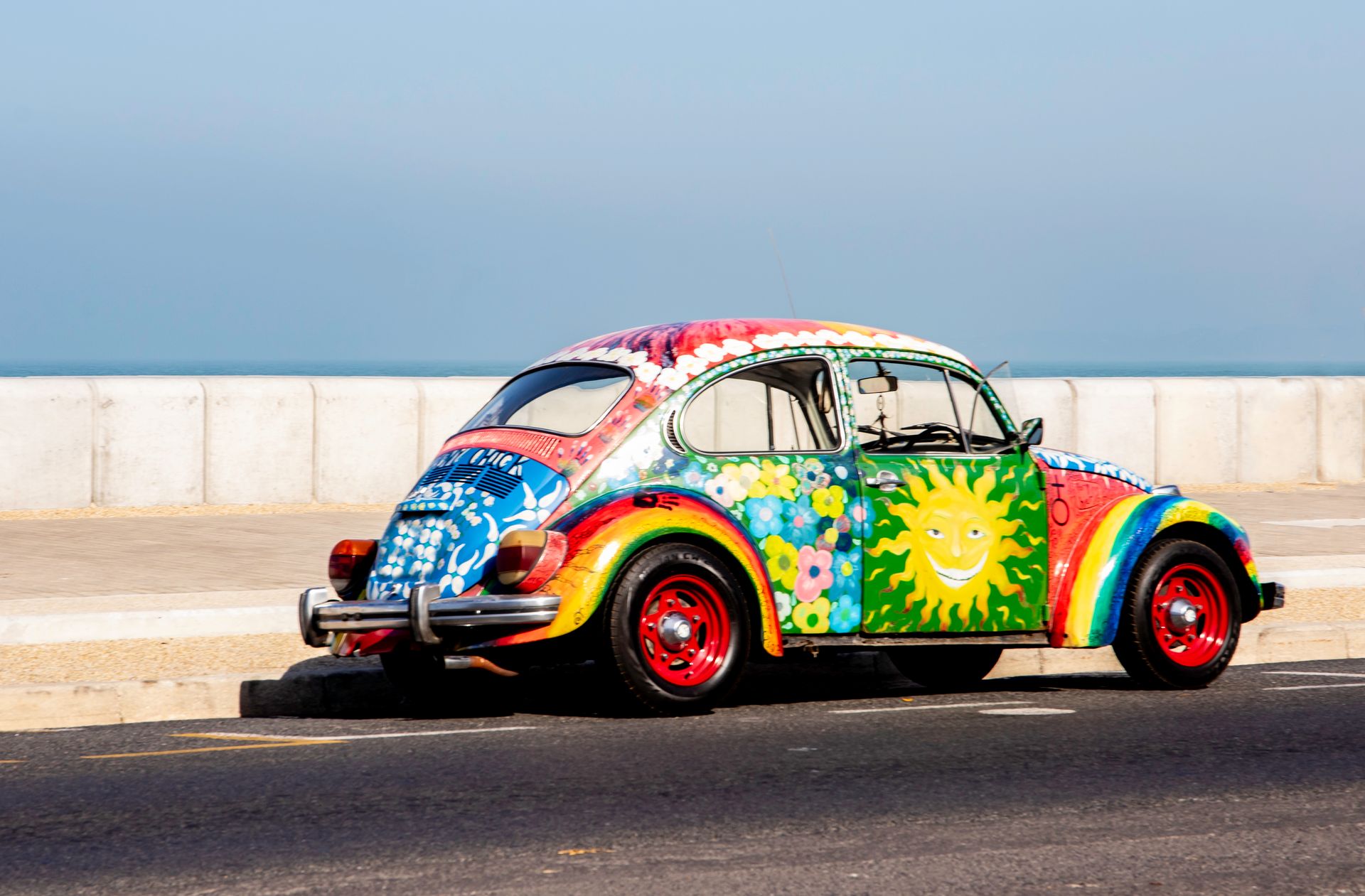 A colorful vintage Volkswagen Beetle with artistic paintwork parked by the ocean under a clear sky.