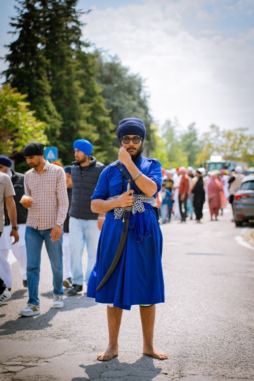 Free A Sikh man dressed in traditional blue attire holds a sword at an outdoor event with multiple people. Stock Photo