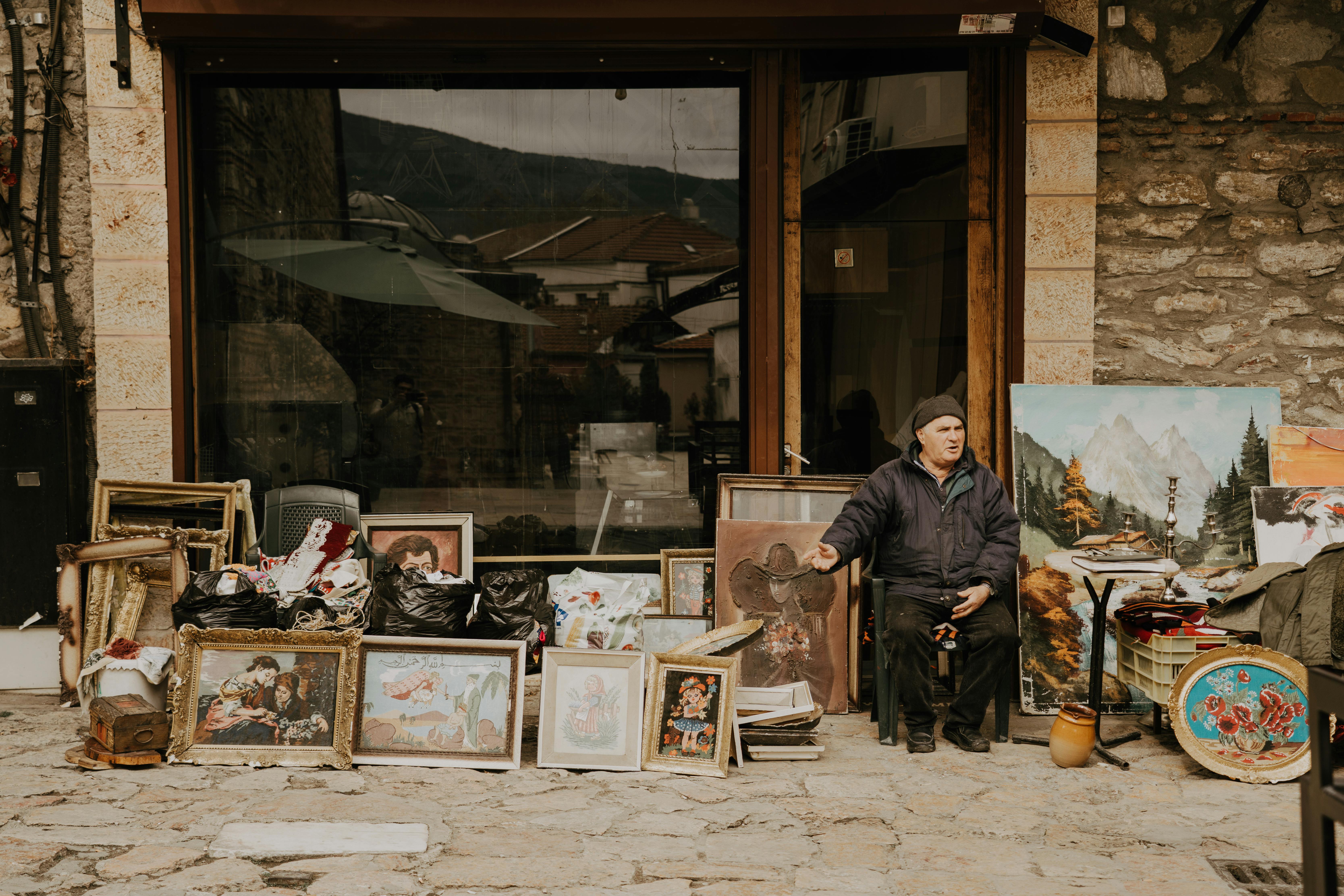 A street vendor sits beside a colorful collection of vintage paintings and art frames outside a rustic shop.