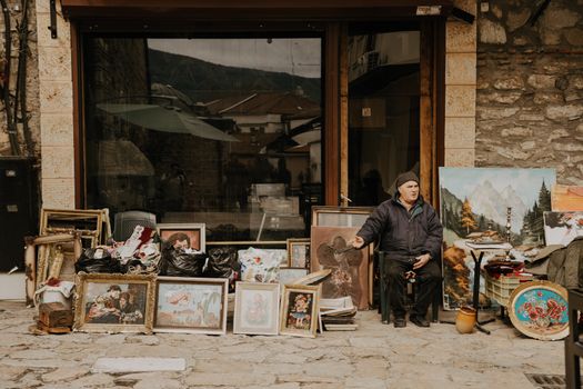 A street vendor sits beside a colorful collection of vintage paintings and art frames outside a rustic shop.