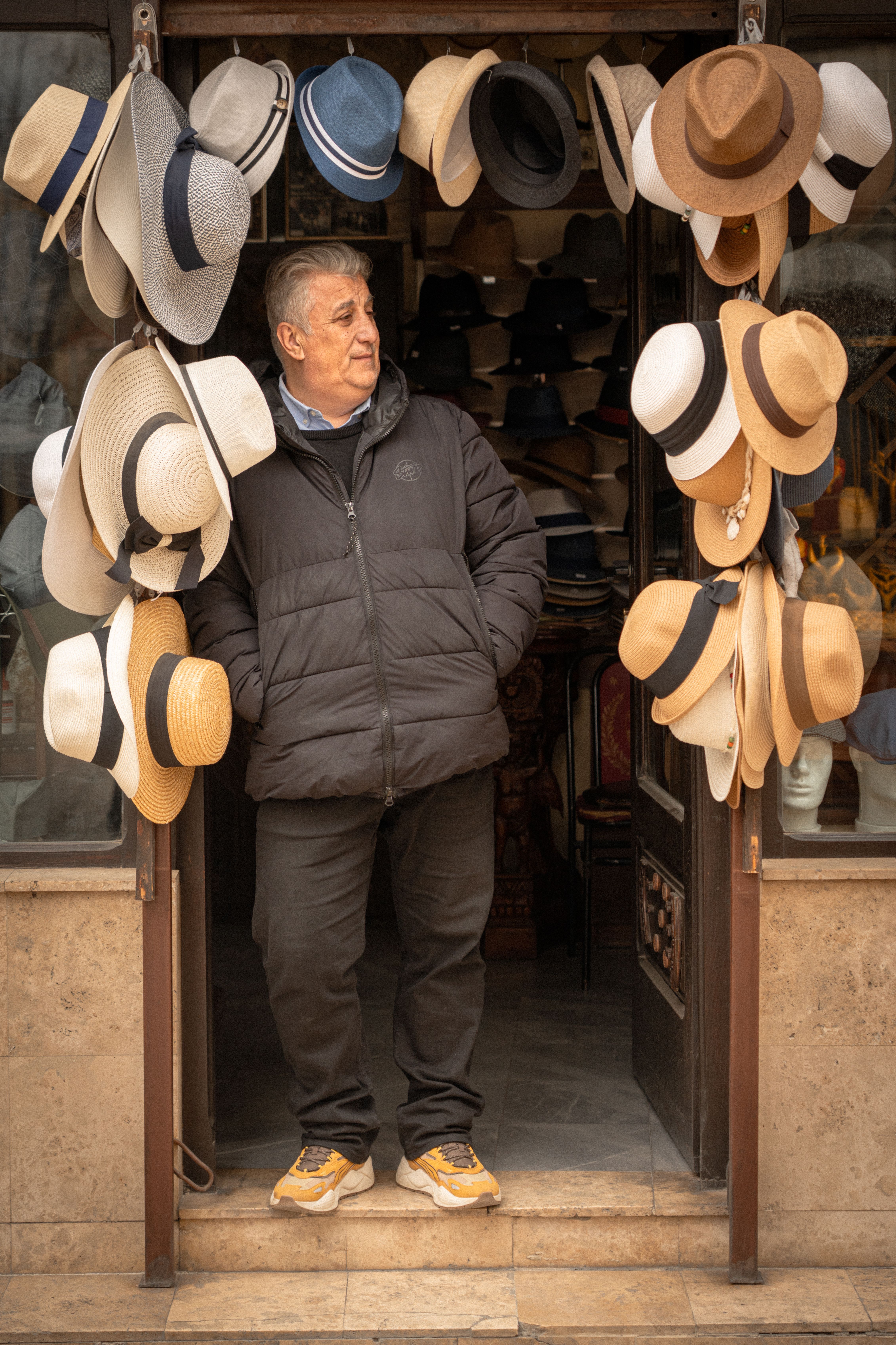 An adult man stands in a hat shop doorway, surrounded by various stylish hats on display.