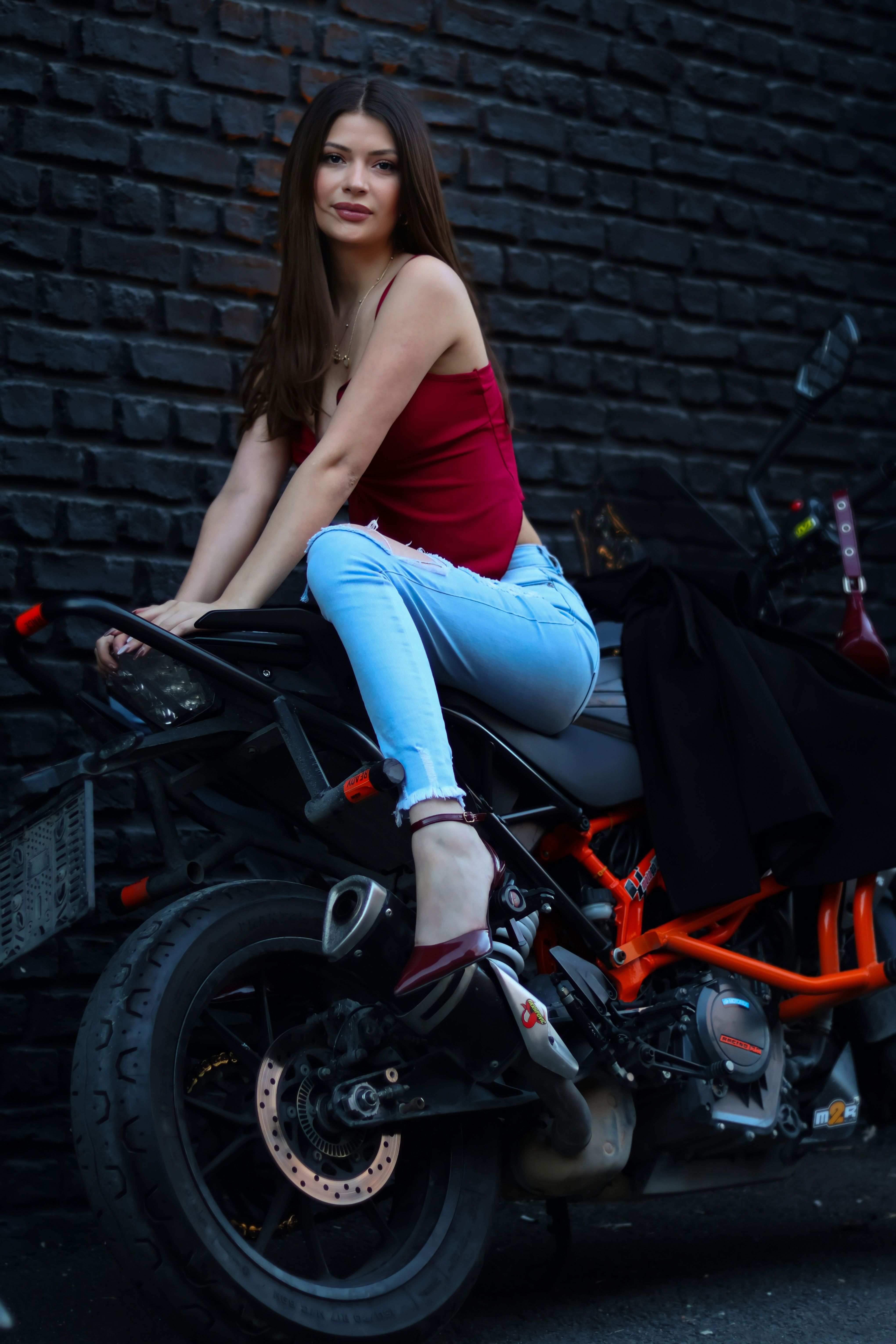 Fashionable woman in red top and jeans seated on a stylish motorcycle against a black brick wall.