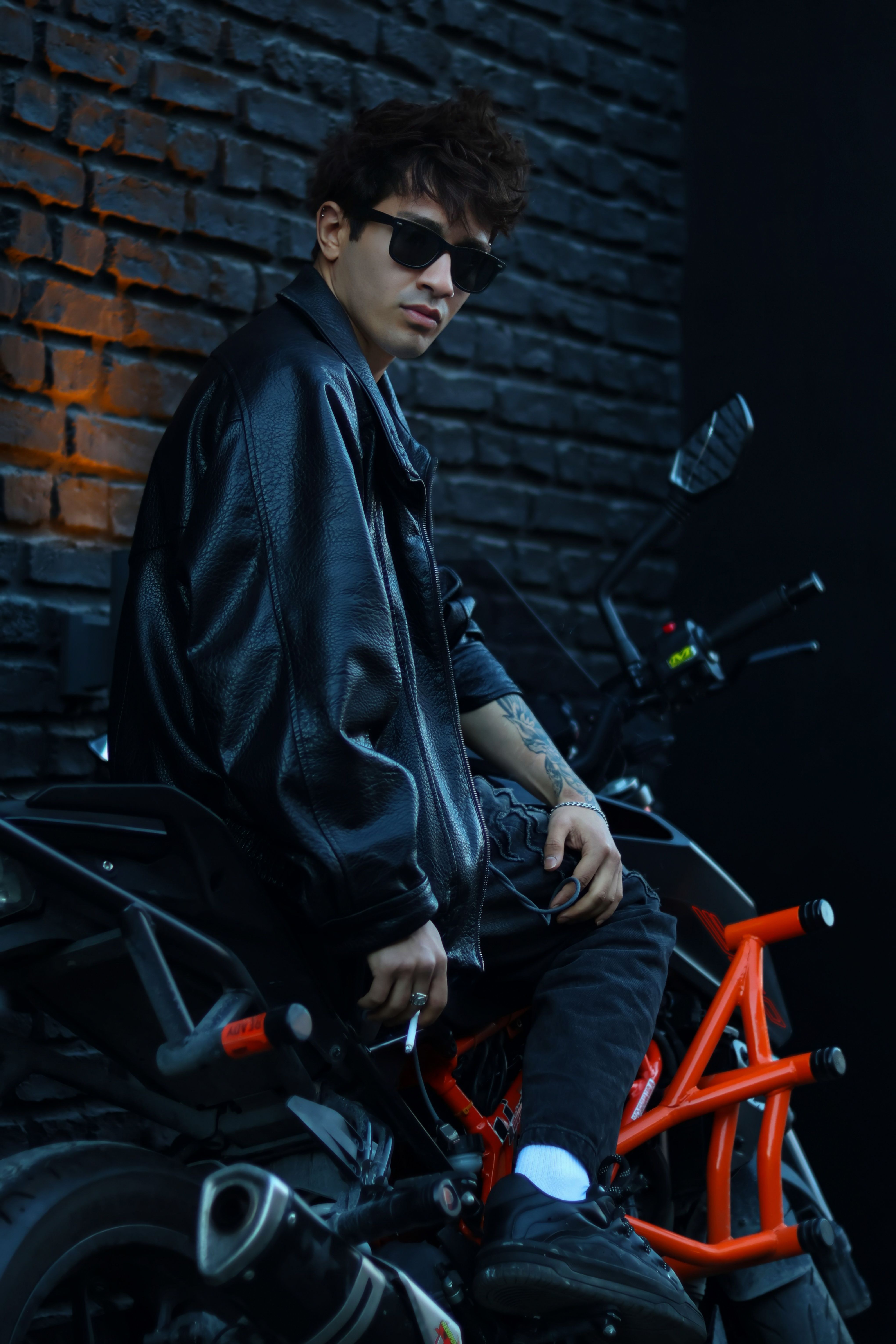 Stylish young man on a motorcycle, wearing sunglasses and a leather jacket against a brick wall.