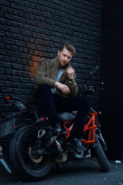Stylish man in a jacket sitting on a motorcycle beside a dark brick wall
