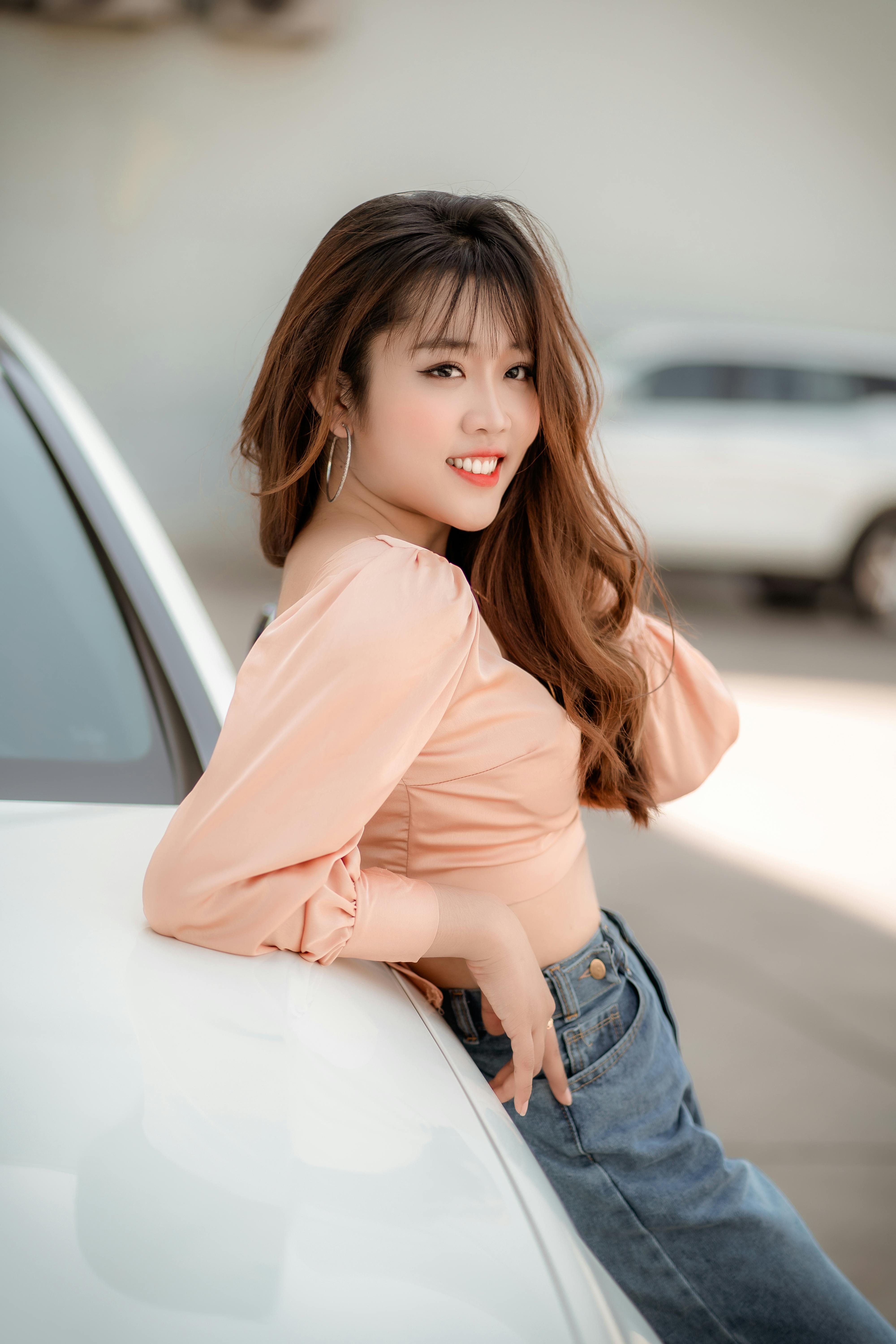 A young woman with long brown hair and orange lipstick leans casually on a car outdoors.