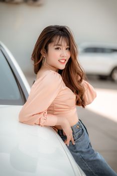 一名年轻女子随意地靠在户外的一辆汽车上，她留着棕色长发，涂着橙色口红