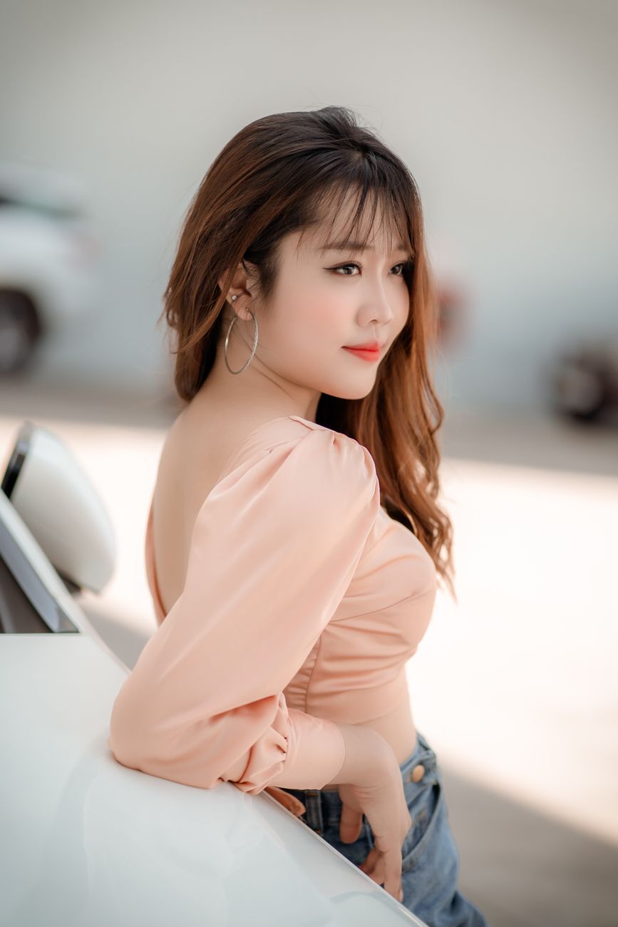 Side view young content Asian lady in stylish outfit leaning on white car motor hood and looking awa