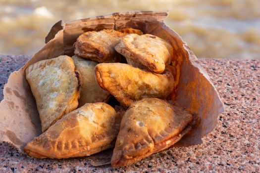 Brown paper bag filled with golden, crispy empanadas sitting on a stone surface by the Montevideo seaside.