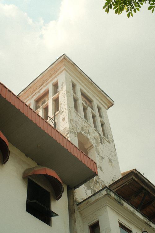 Free A historic building with a tall tower under a cloudy sky, featuring a red roof and worn exterior. Stock Photo