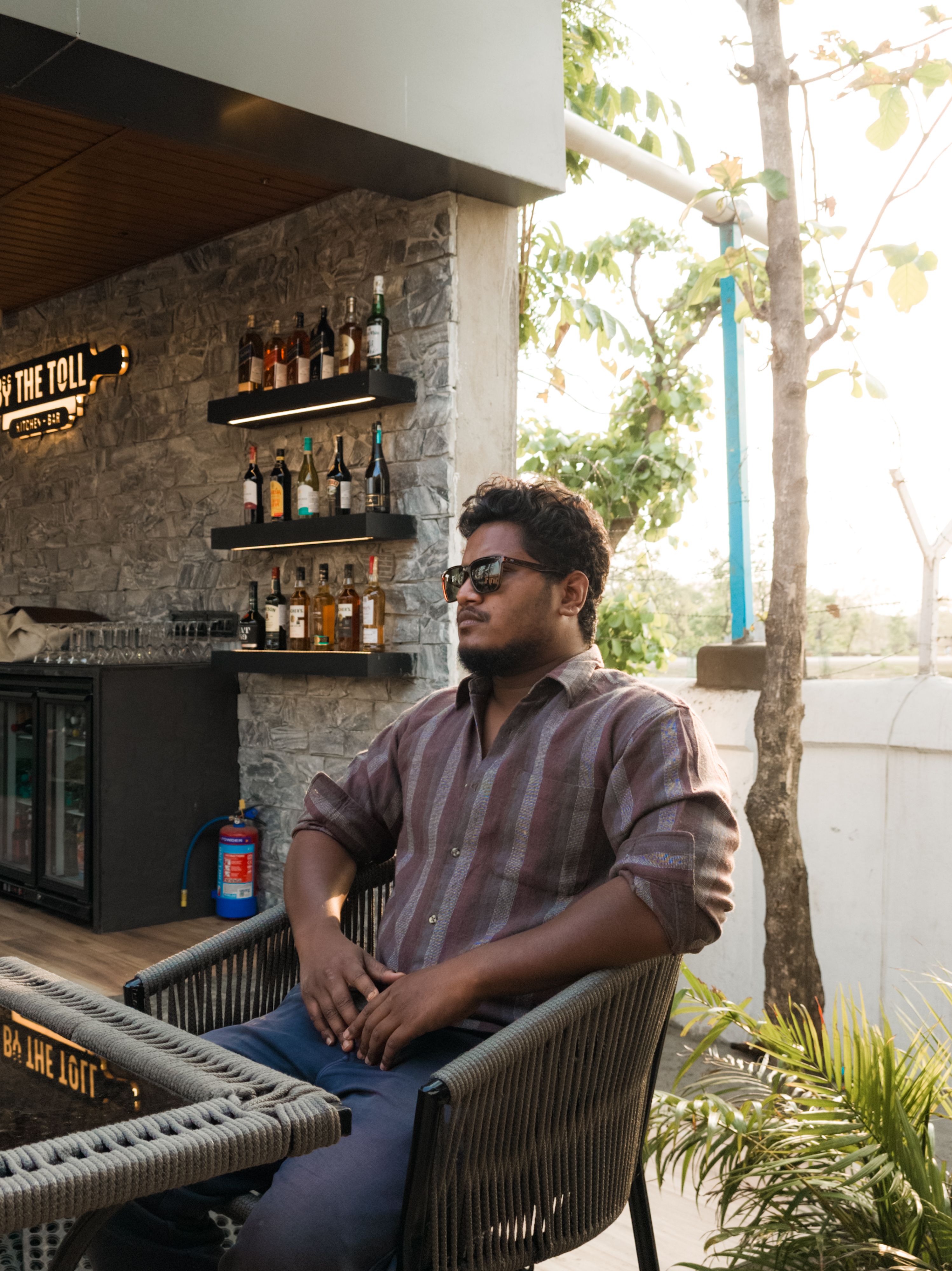 Man in sunglasses relaxing at an outdoor cafe bar surrounded by bottles in daylight.