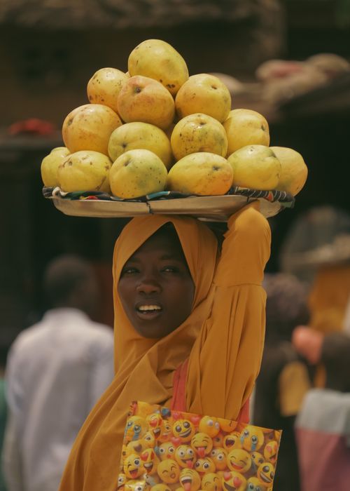 Free A woman in colorful attire balances a tray of mangoes on her head in a bustling market setting. Stock Photo