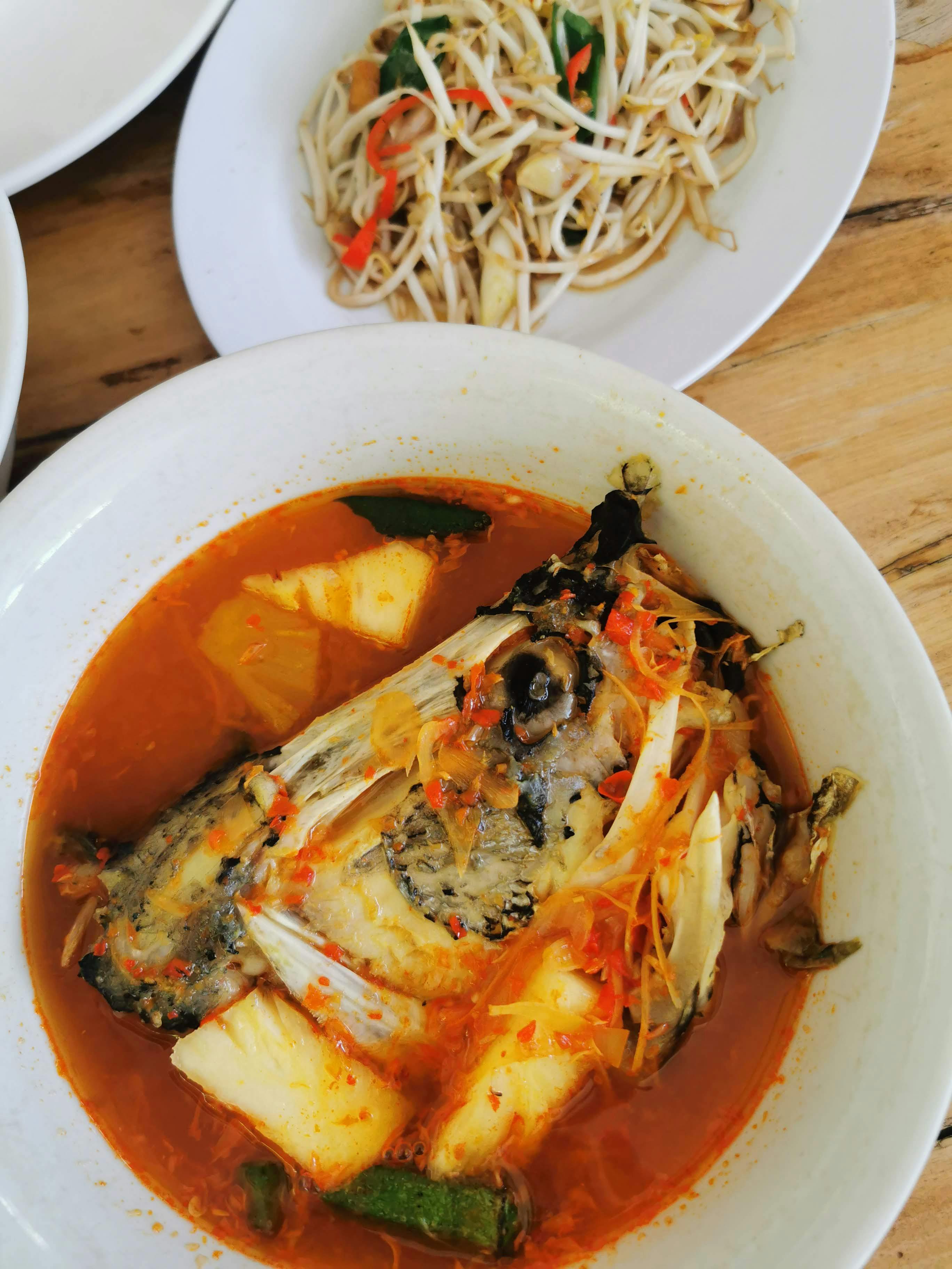 A bowl of vibrant orange Thai sour curry with chunks of white fish, fresh pineapple, and tomato wedges floating in an aromatic broth, served alongside a mound of jasmine rice