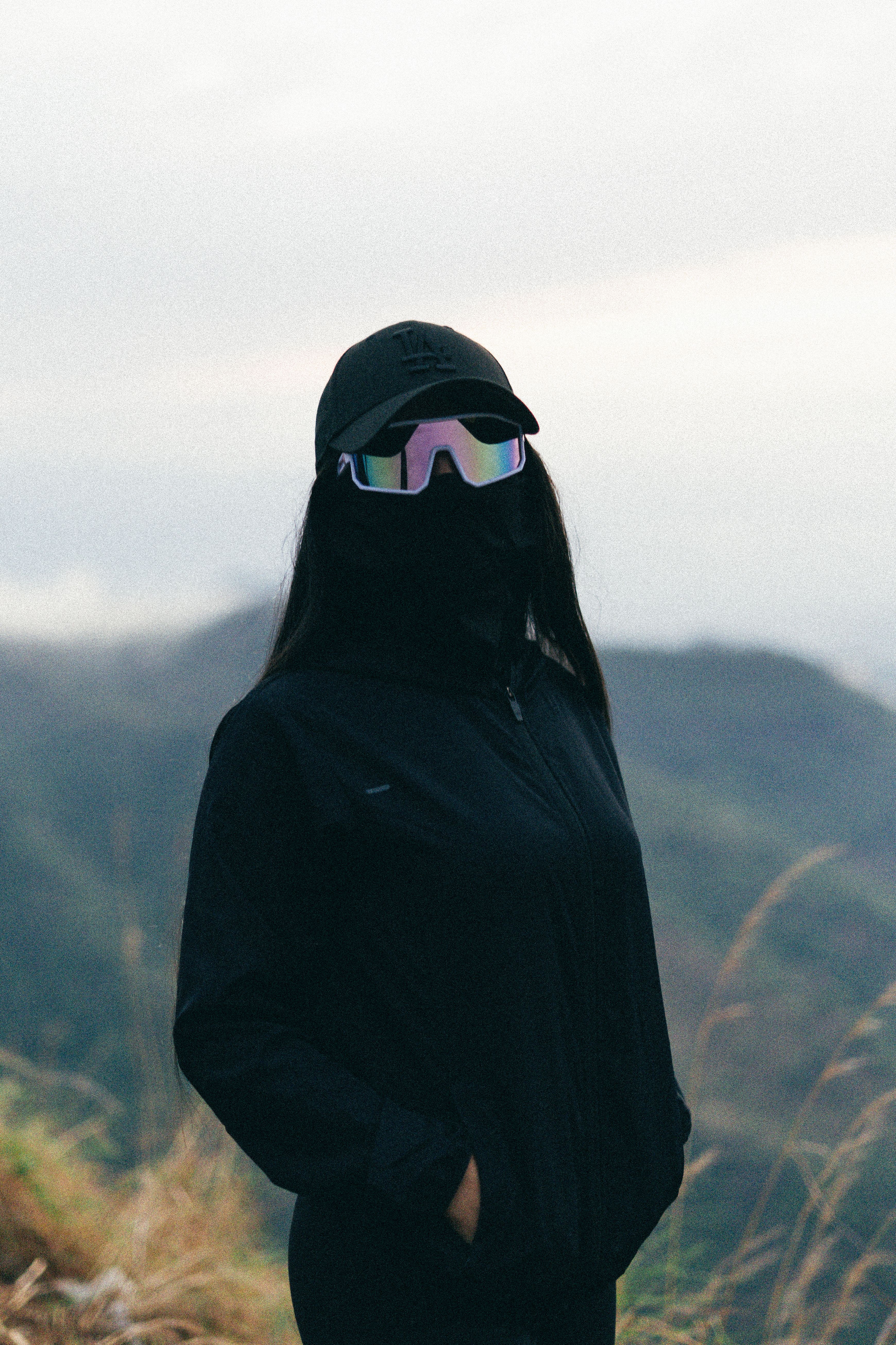 A woman in black with reflective sunglasses stands in a mountainous landscape, exuding mystery.