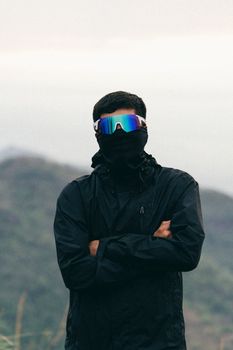 An individual wearing a mask and black jacket outdoors, with a mountainous backdrop.