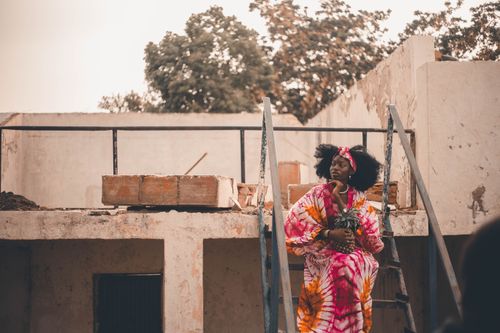 Free A woman sits on a rooftop wearing a colorful tie-dye dress, surrounded by a rustic setting. Stock Photo