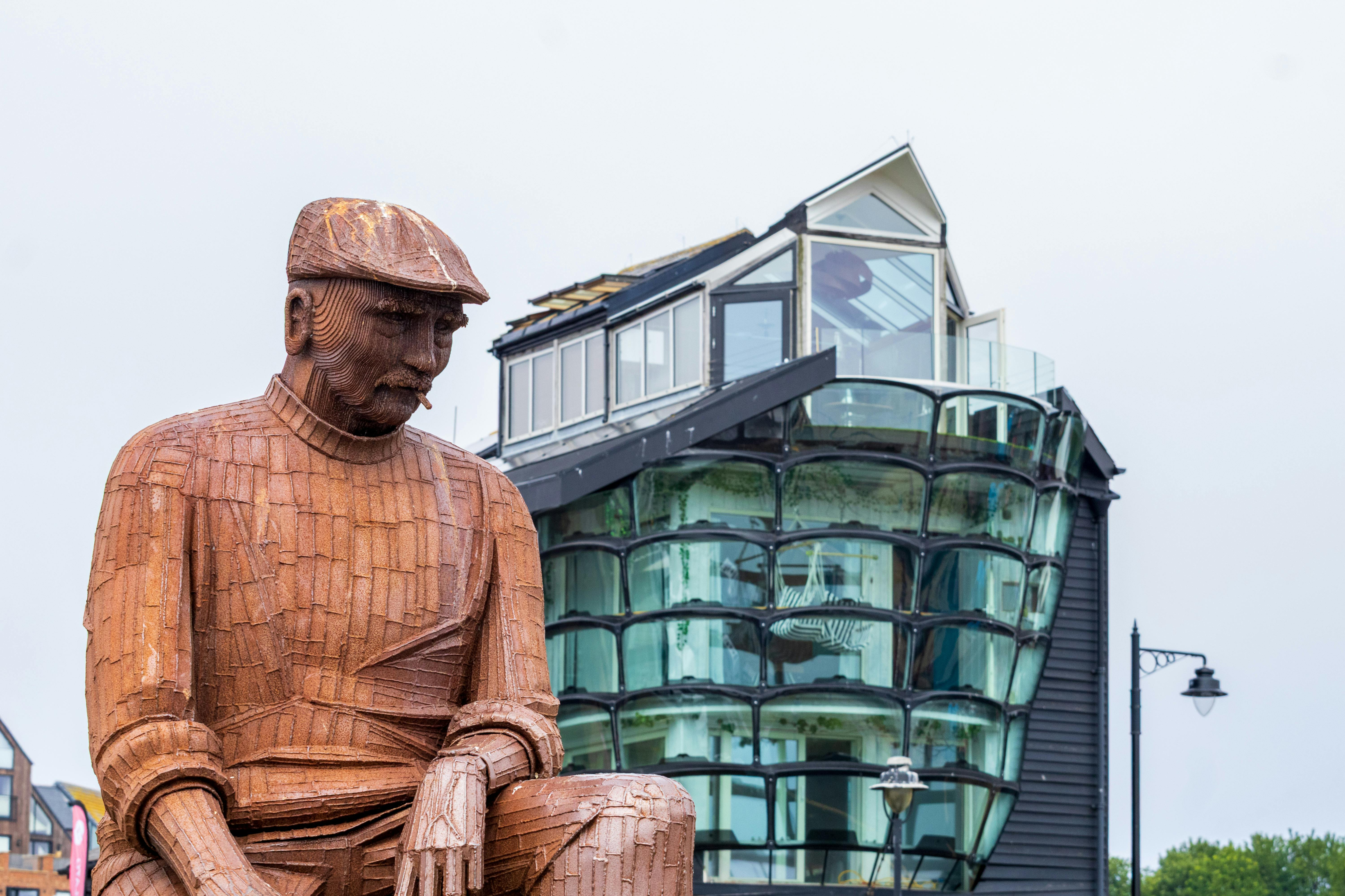 A wooden statue of a man sits near a unique modern glass building against a cloudy sky.