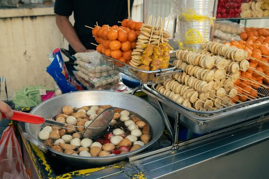 Street food vendor cooking skewered snacks and dumplings in a large pan at an outdoor market.