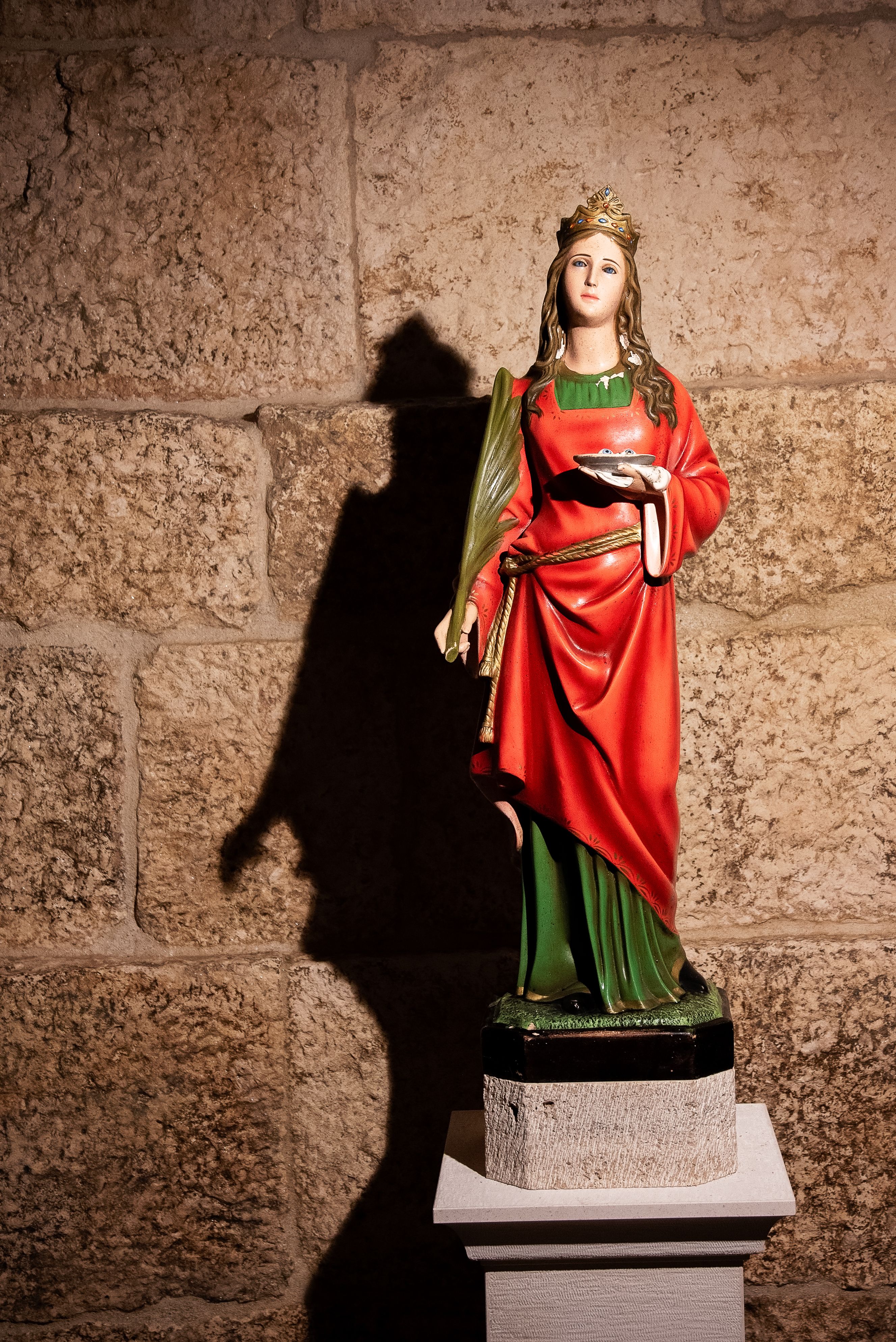A statue of a saint against a stone wall with dramatic lighting casting shadows.