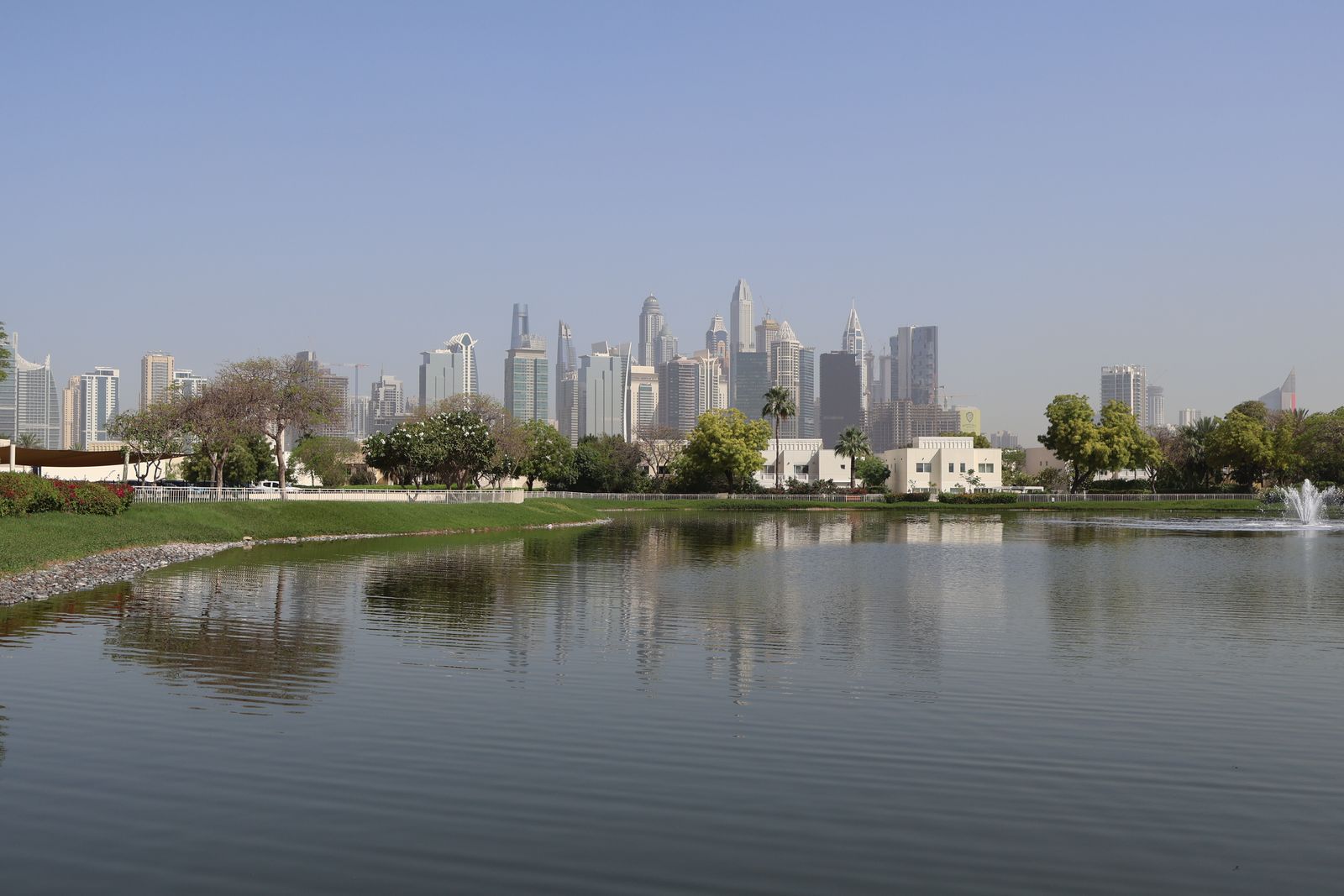 Community lake with fountains, green banks and low-rise homes below the Dubai skyline