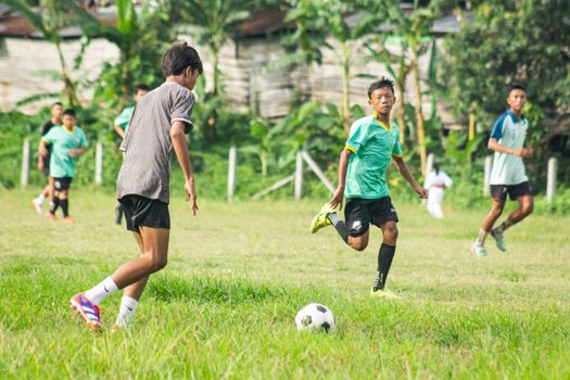 Teenagers playing soccer on a grassy field in Myawaddy, Myanmar on a sunny day.