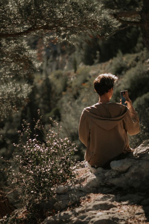 Free A young man takes a photo with his phone while sitting on a rocky ledge in a lush forest. Stock Photo