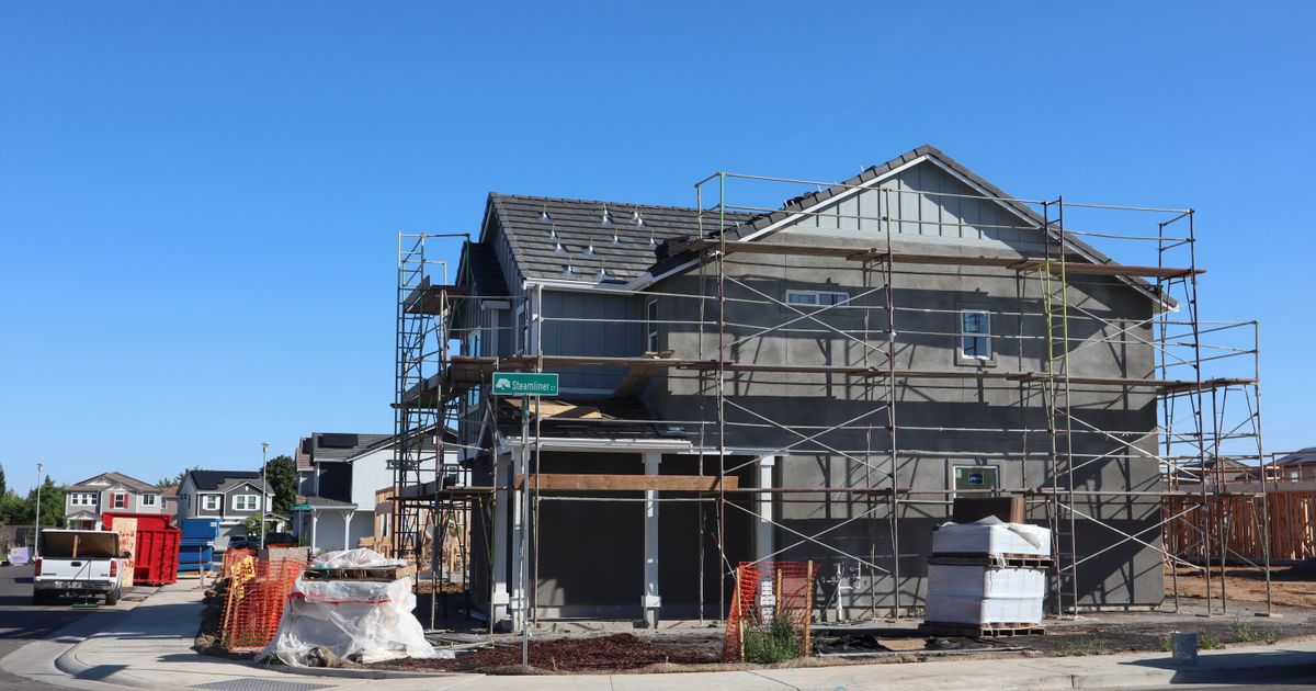 A new home under construction behind scaffolding on a corner lot, with finished houses visible down the street.