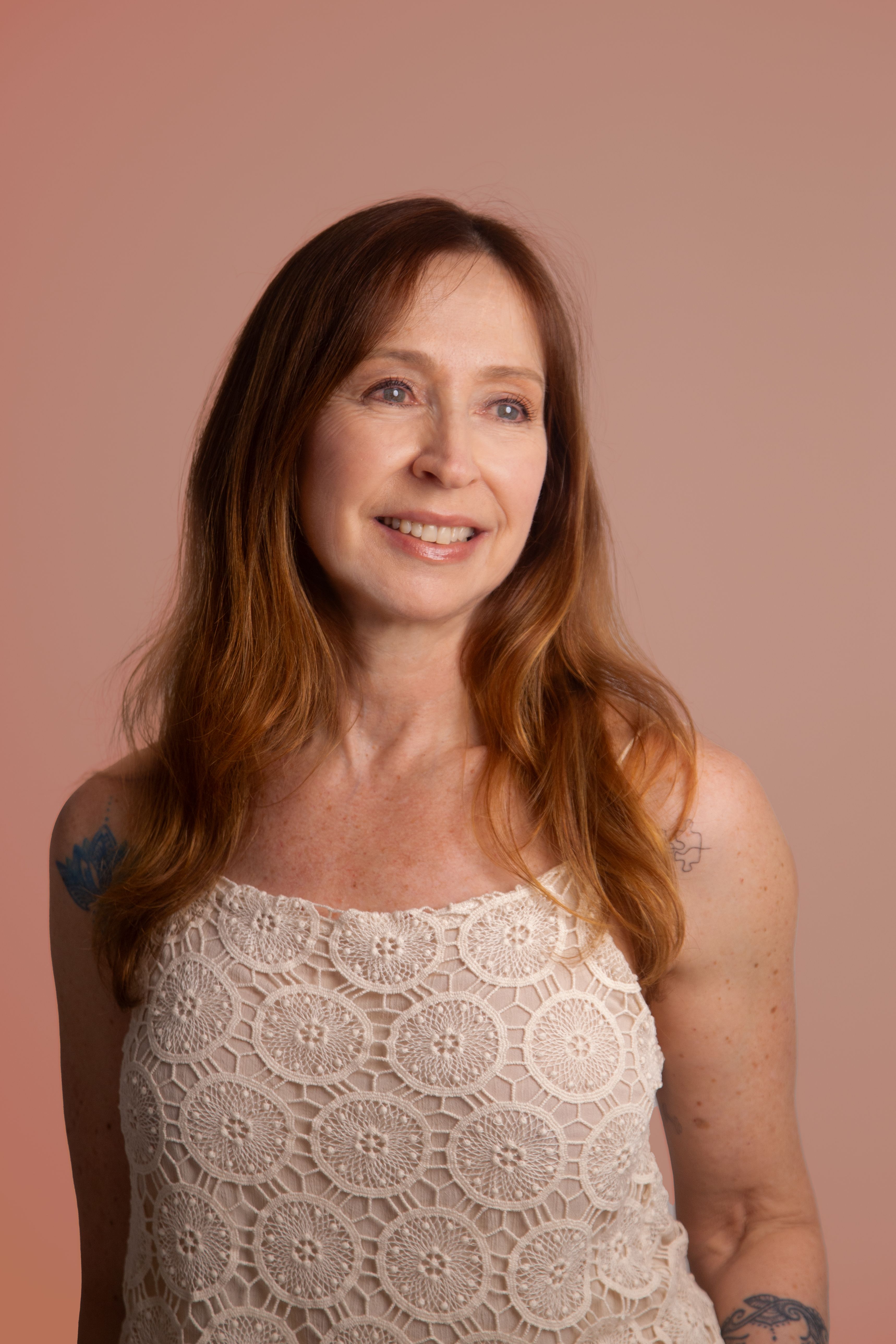 A woman with long hair and tattoos smiles warmly against a neutral backdrop.