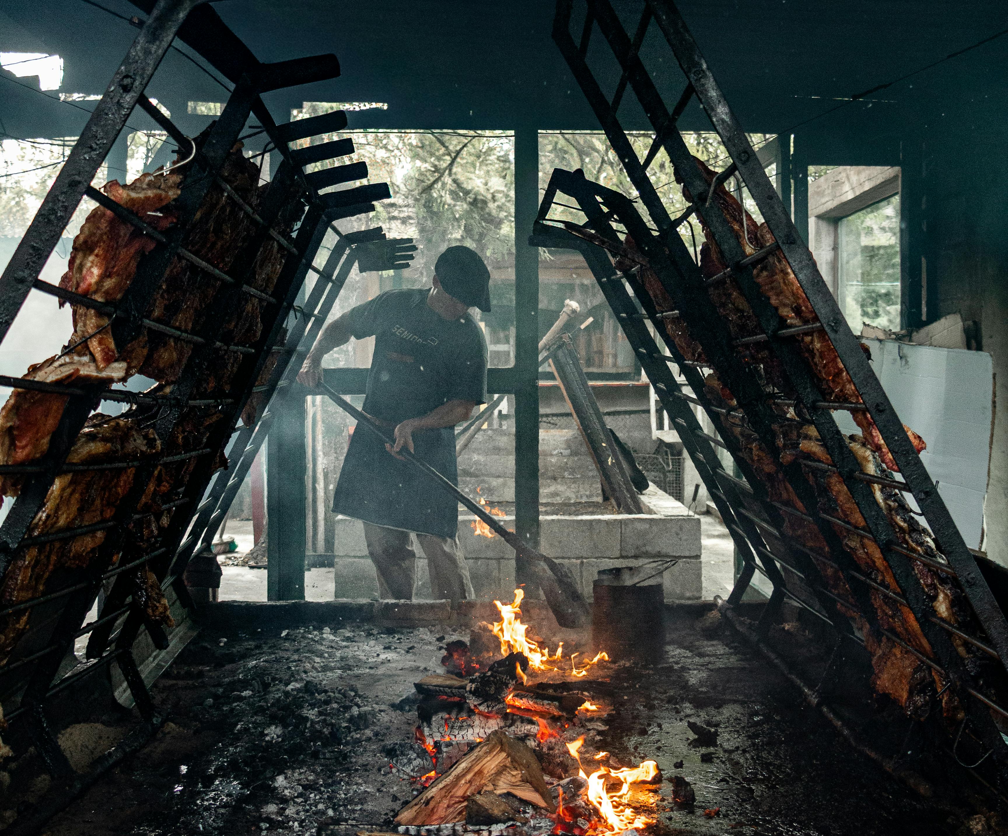 Traditional asado barbecue with a person tending to meat on an open fire grill.
