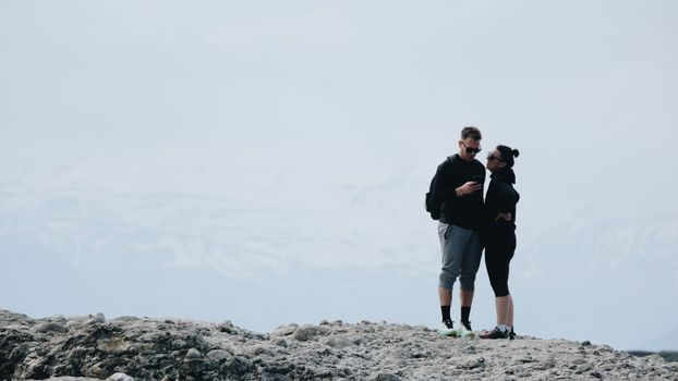 A couple enjoys a moment of embrace on a rocky mountain peak with snow-capped backgrounds.