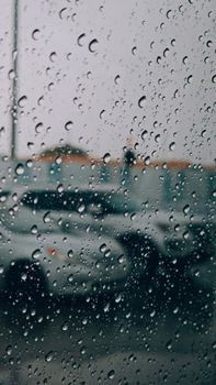 Close-up view of raindrops on glass with blurry cars in a parking lot background.