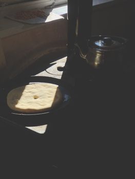 A flatbread cooking on a stove next to a metal teapot in a dimly lit kitchen.