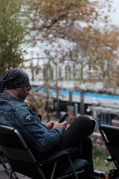 Casual man with headscarf using smartphone outside while sitting in a relaxed position.